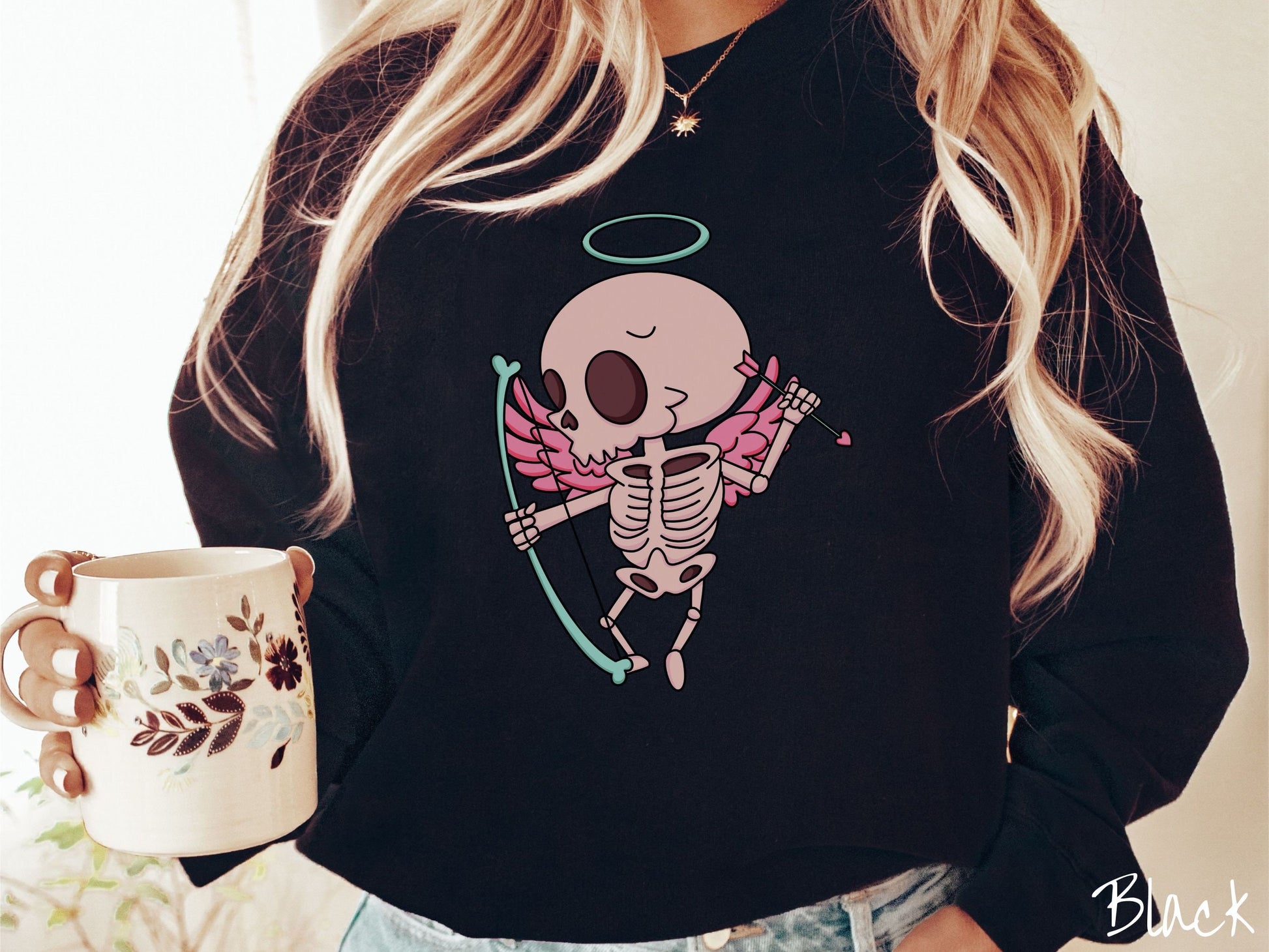 A woman wearing a cute black colored sweatshirt with a pink winged baby skeleton cupid with a halo holding a heart-tipped arrow in its left hand and a bow in its right hand.