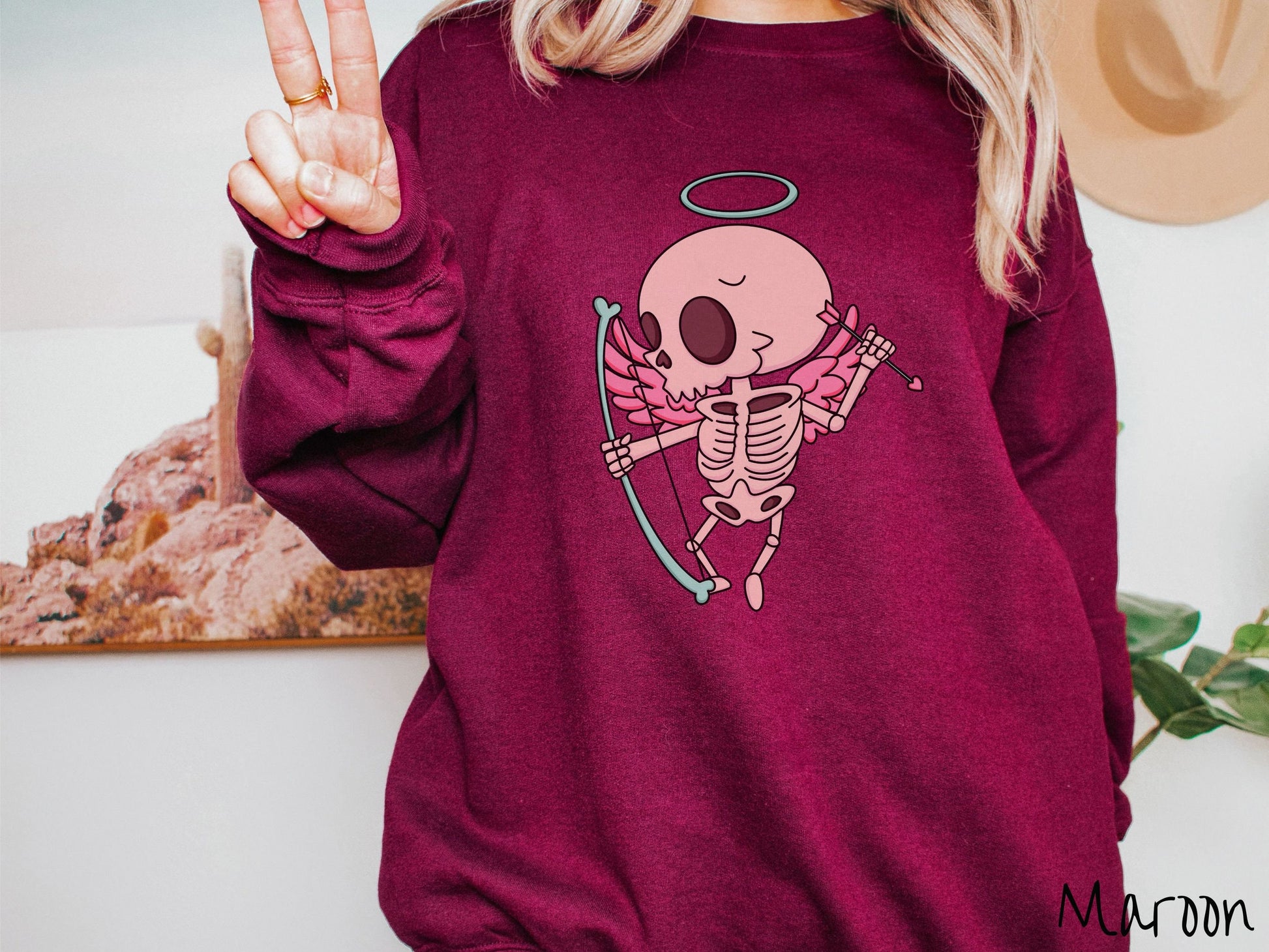 A woman wearing a cute maroon colored sweatshirt with a pink winged baby skeleton cupid with a halo holding a heart-tipped arrow in its left hand and a bow in its right hand.