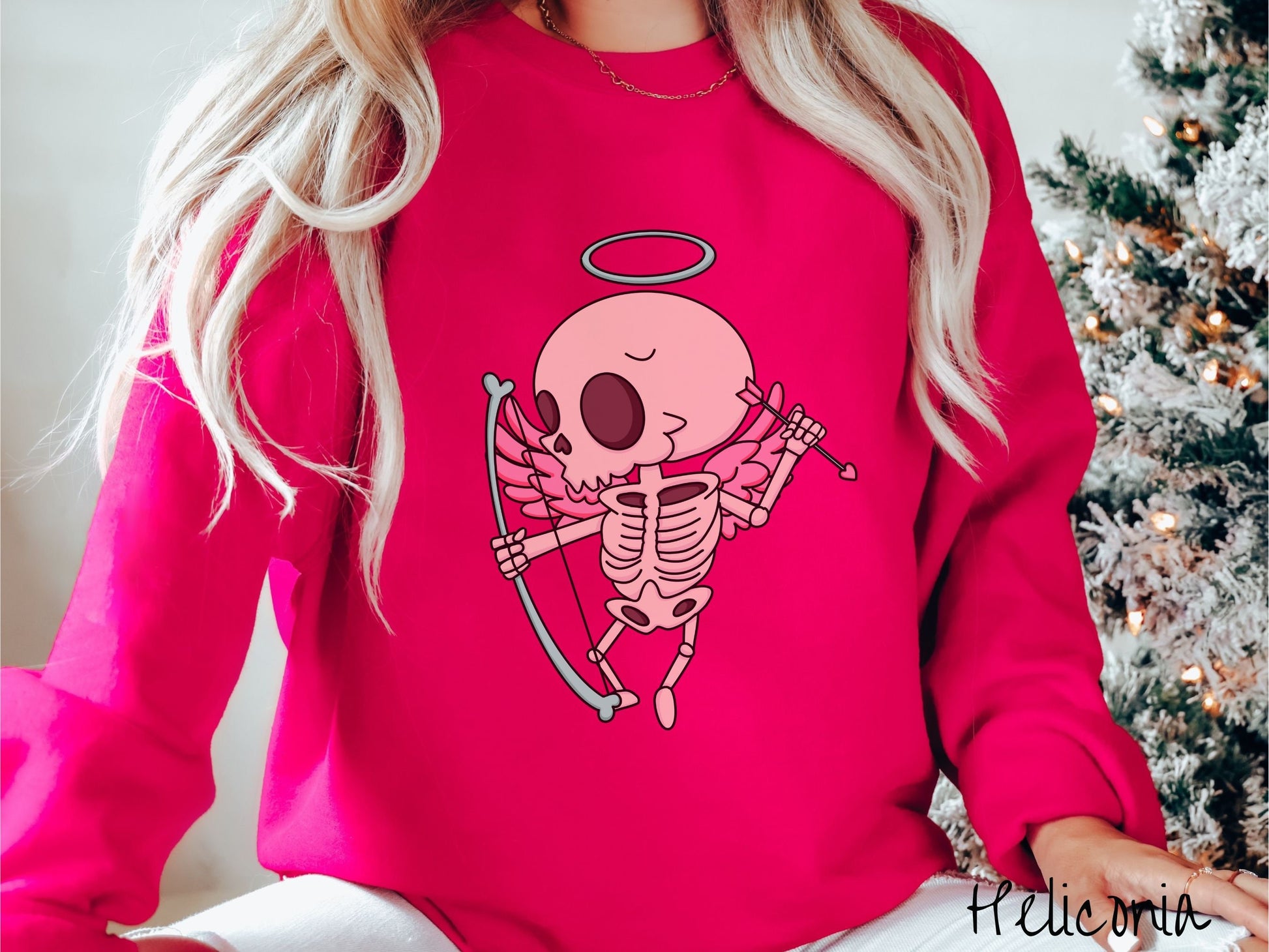 A woman wearing a cute heliconia colored sweatshirt with a pink winged baby skeleton cupid with a halo holding a heart-tipped arrow in its left hand and a bow in its right hand.