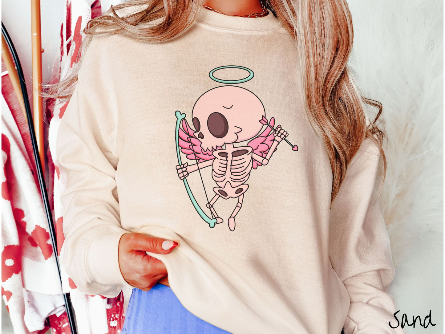 A woman wearing a cute sand colored sweatshirt with a pink winged baby skeleton cupid with a halo holding a heart-tipped arrow in its left hand and a bow in its right hand.