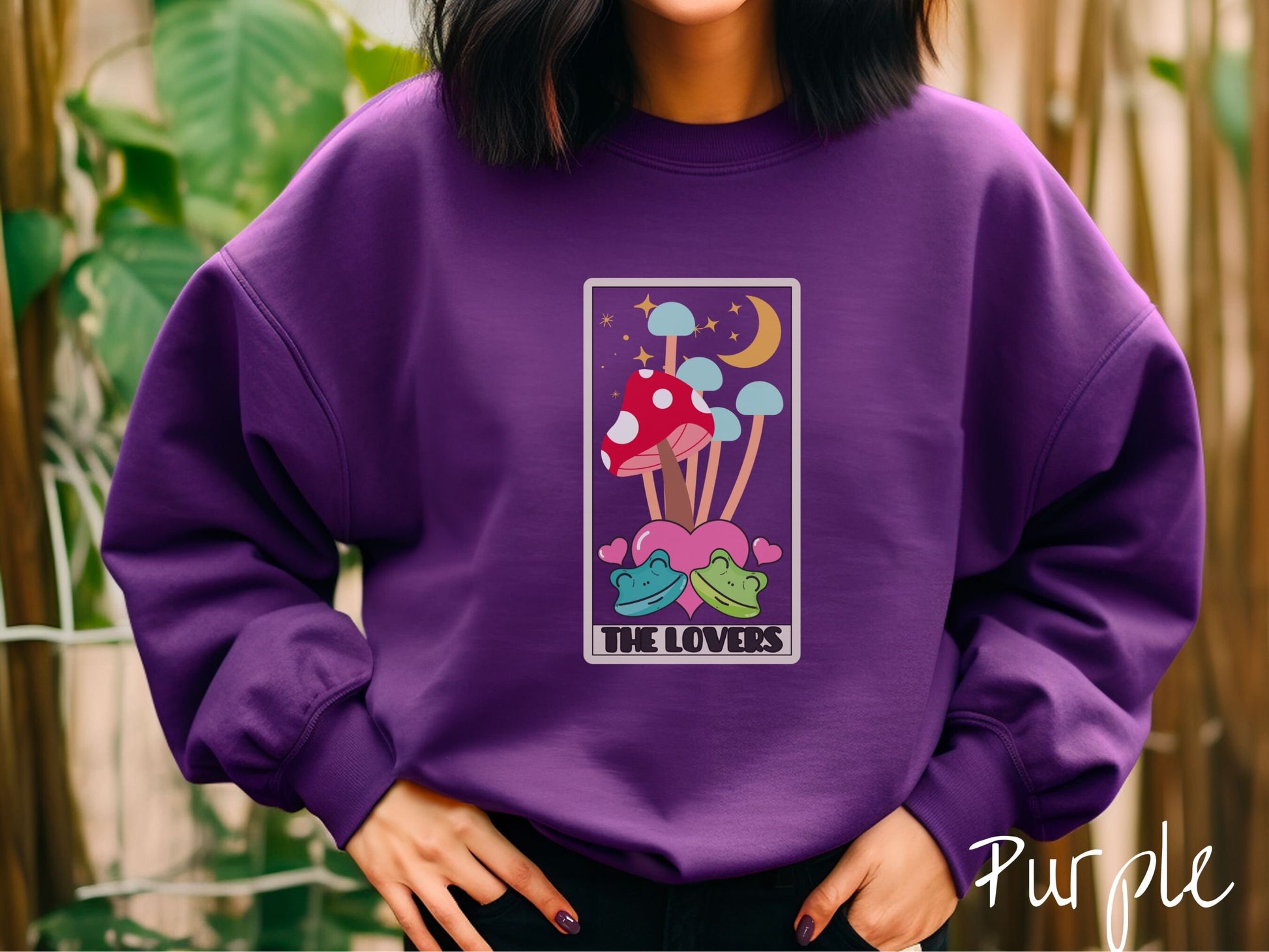 A woman wearing a cute purple colored sweatshirt with a large red mushroom and 4 turquoise mushrooms in the background, all under a crescent yellow moon and yellow stars, two green frogs smiling in front of pink hearts below the mushrooms.