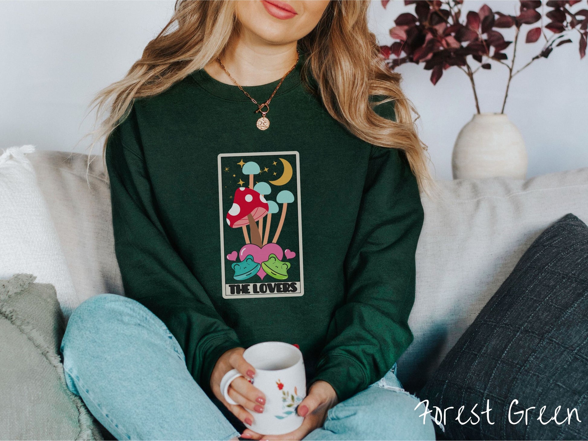 A woman wearing a cute forest green colored sweatshirt with a large red mushroom and 4 turquoise mushrooms in the background, all under a crescent yellow moon and yellow stars, two green frogs smiling in front of pink hearts below the mushrooms.
