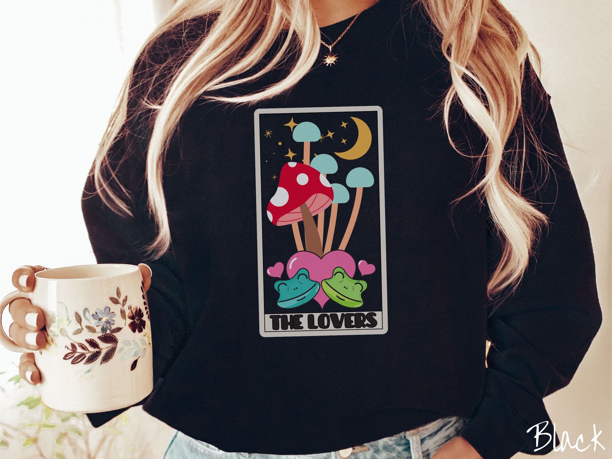 A woman wearing a cute black colored sweatshirt with a large red mushroom and 4 turquoise mushrooms in the background, all under a crescent yellow moon and yellow stars, two green frogs smiling in front of pink hearts below the mushrooms.
