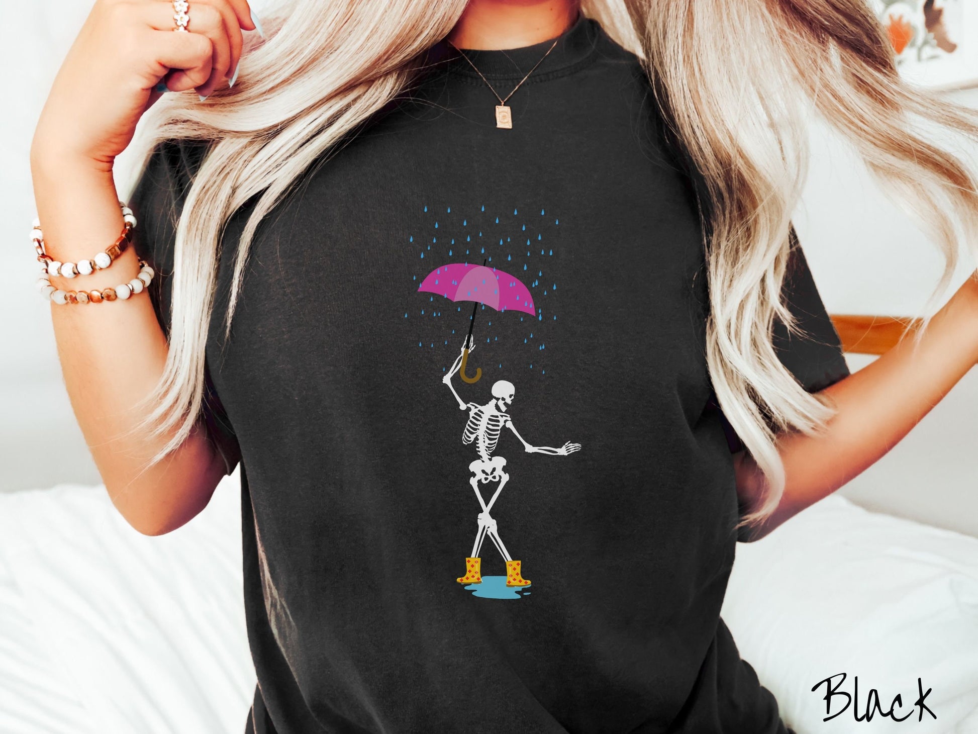 A woman wearing a cute black colored shirt with a white female skeleton dancing in the rain with a pink and purple umbrella and wearing yellow with red polka dot rain boots.