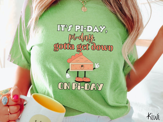 A woman wearing a cute, vintage kiwi colored comfort colors shirt with text Its Pi-day, pi-day, gotta get down on pi-day in white and orange font. There is a smiling piece of pumpkin pie with arms with white gloves and legs with orange shoes on.