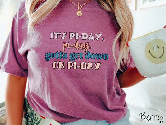 A woman wearing a cute, vintage berry colored comfort colors shirt with the text It&#39;s Pi-day, pi-day, gotta get down on pi-day in white, blue, and orange font.