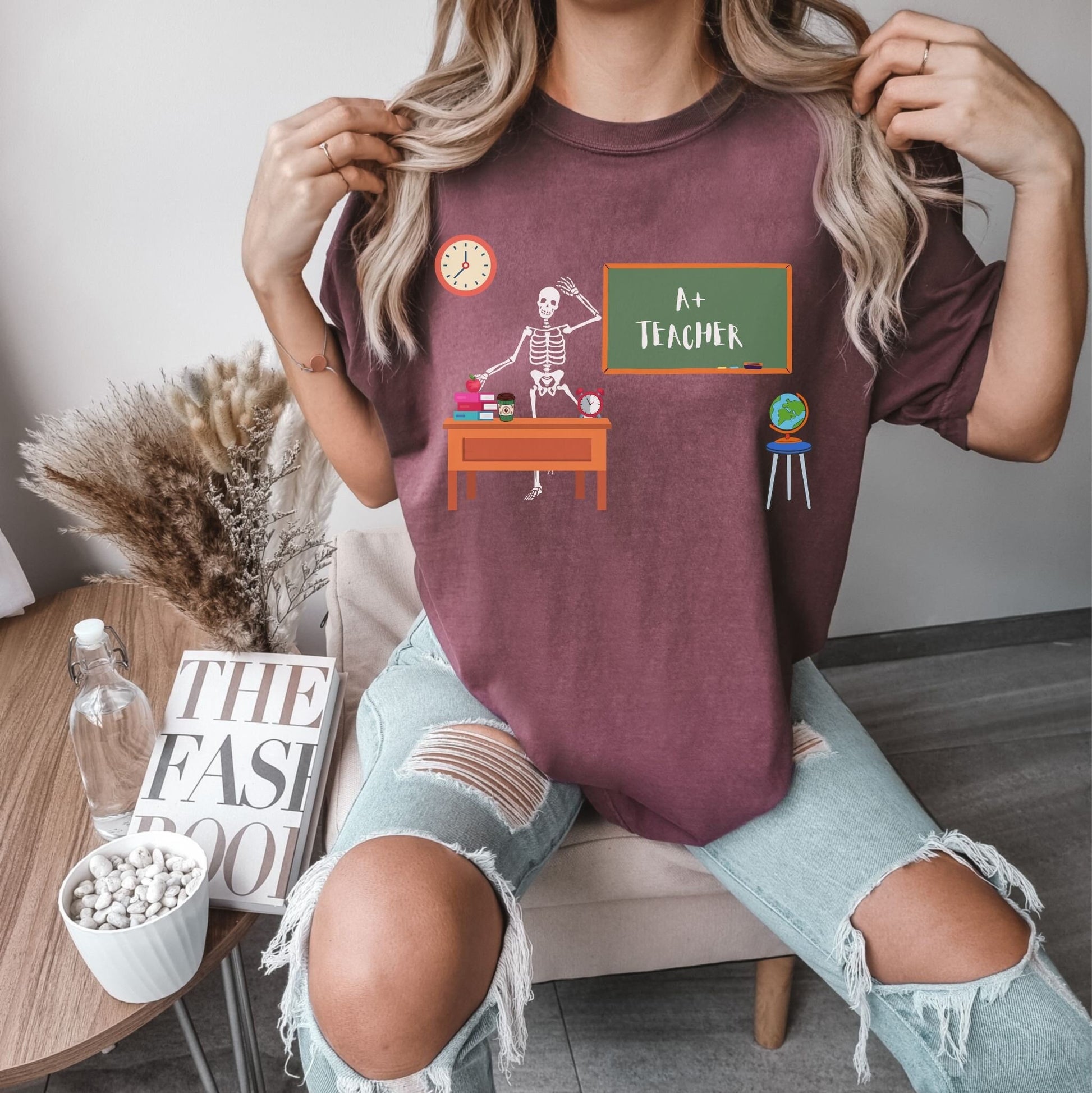 A woman wearing a cute, vintage vineyard colored shirt with a skeleton teacher waving from their desk in front of the green chalkboard which has written A+ Teacher on it. On the teacher’s desk are colorful books, an apple, a coffee, and a clock.