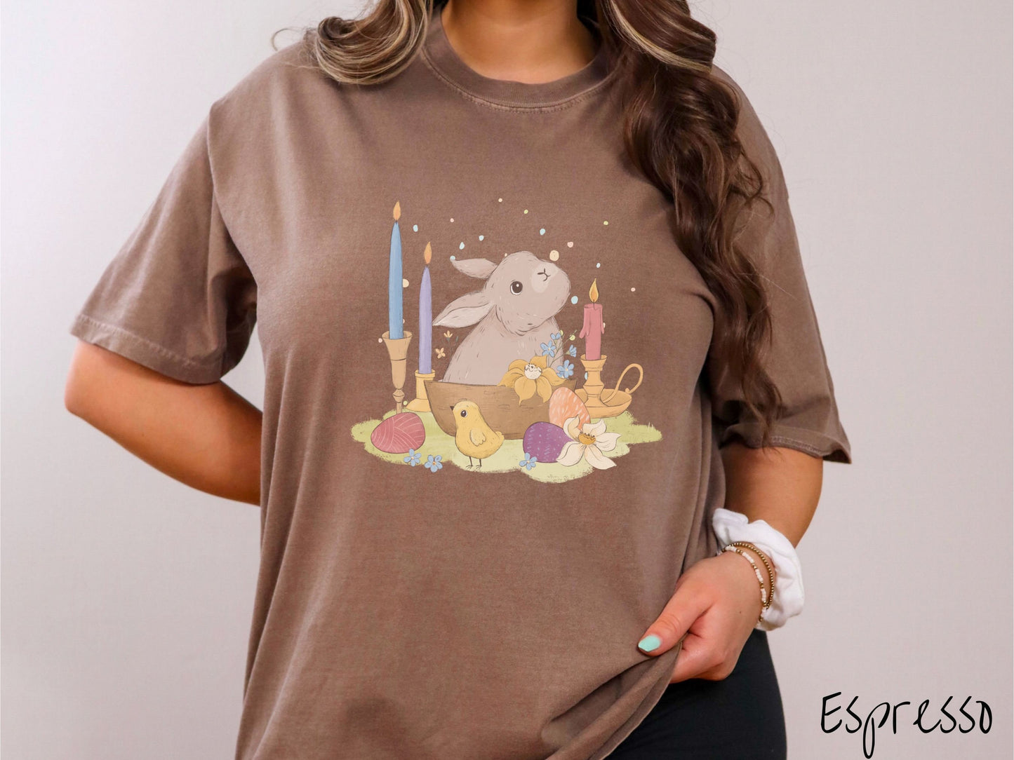 A woman wearing a cute, vintage espresso colored shirt with a gray bunny rabbit sitting in between three lit candles, some red, orange, and purple eggs, and yellow and white flowers.