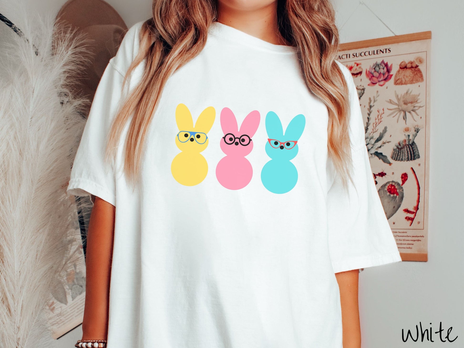 A woman wearing a cute, vintage white colored shirt with three peep-like rabbits on the front. One is yellow and wearing blue glasses, the middle is pink with black glasses, and the one on the right is light blue with red glasses.