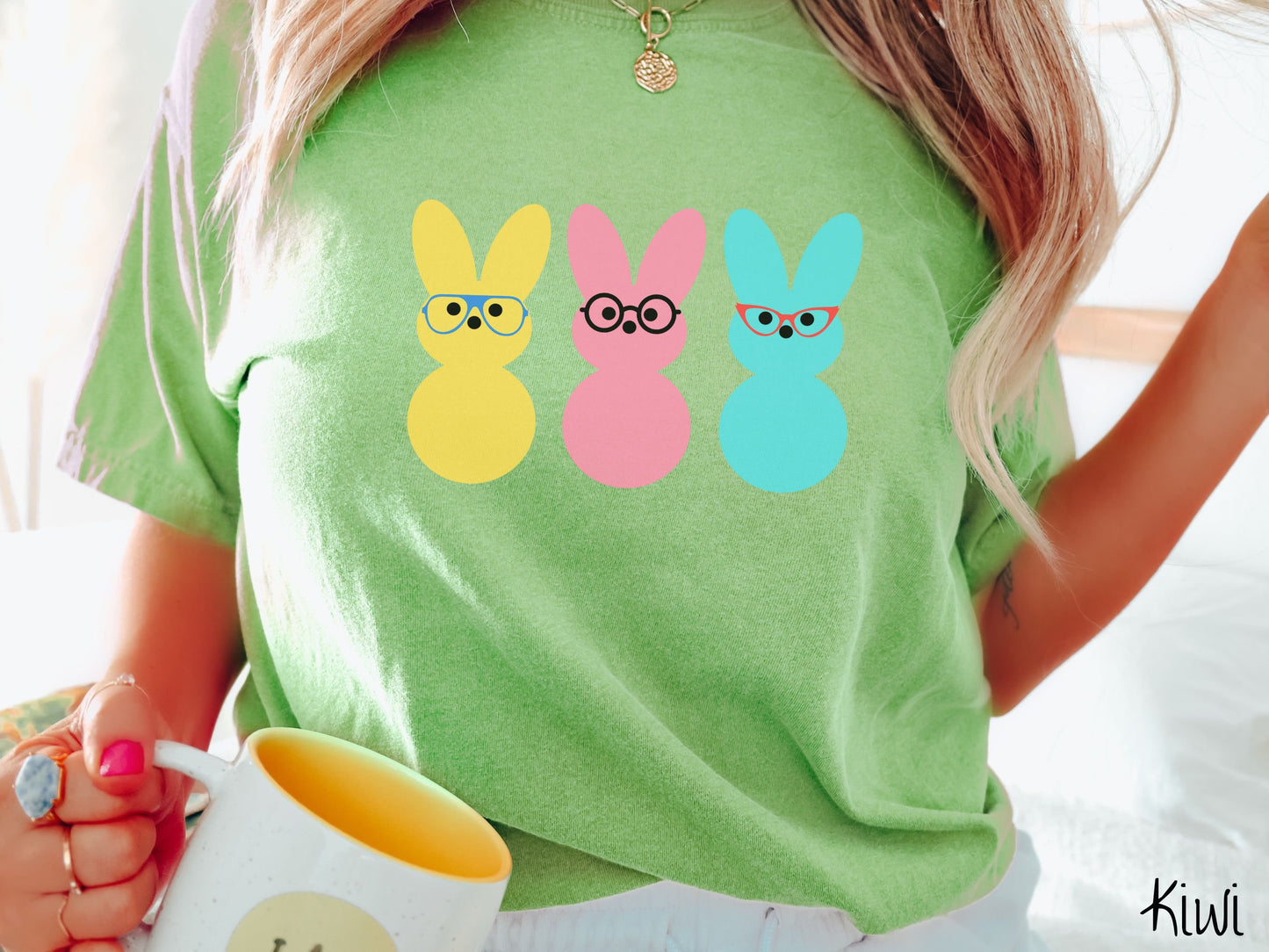 A woman wearing a cute, vintage kiwi colored shirt with three peep-like rabbits on the front. One is yellow and wearing blue glasses, the middle is pink with black glasses, and the one on the right is light blue with red glasses.