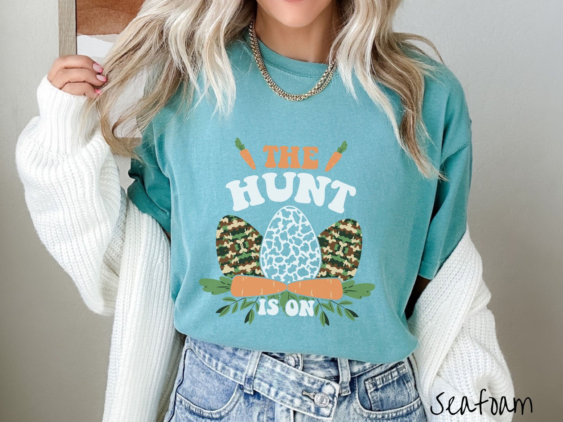 A woman wearing a cute, vintage seafoam colored shirt with the text The Hunt Is On in orange and white font, with three Easter eggs painted in woodland camo and black and white camo in front. Above and below the eggs are carrots with leafy ends.