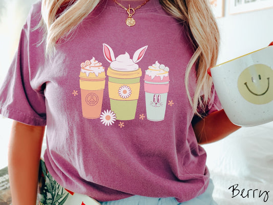 A woman wearing a cute, vintage berry colored shirt with three coffee cups on the front. One is orange with whipped cream and a peace sign. The middle is green with whipped cream, and bunny ears, and the third is light blue with whipped cream.