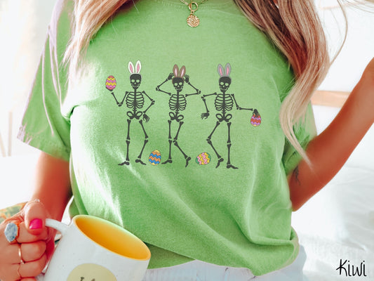 A woman wearing a cute, vintage kiwi colored shirt with three skeletons wearing white, brown, and gray colored bunny ears posing next to each other holding colorful Easter eggs. There are a couple colorful, striped Easter eggs at their feet.