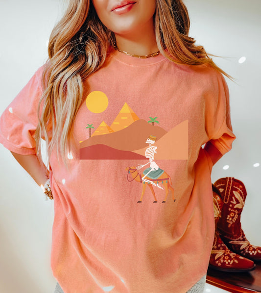 A woman wearing a cute, vintage terracotta colored shirt with a skeleton wearing an Egyptian hat riding a brown camel through the desert, the Egyptian pyramids in the distance behind sand dunes and palm trees with the sun high in the sky.