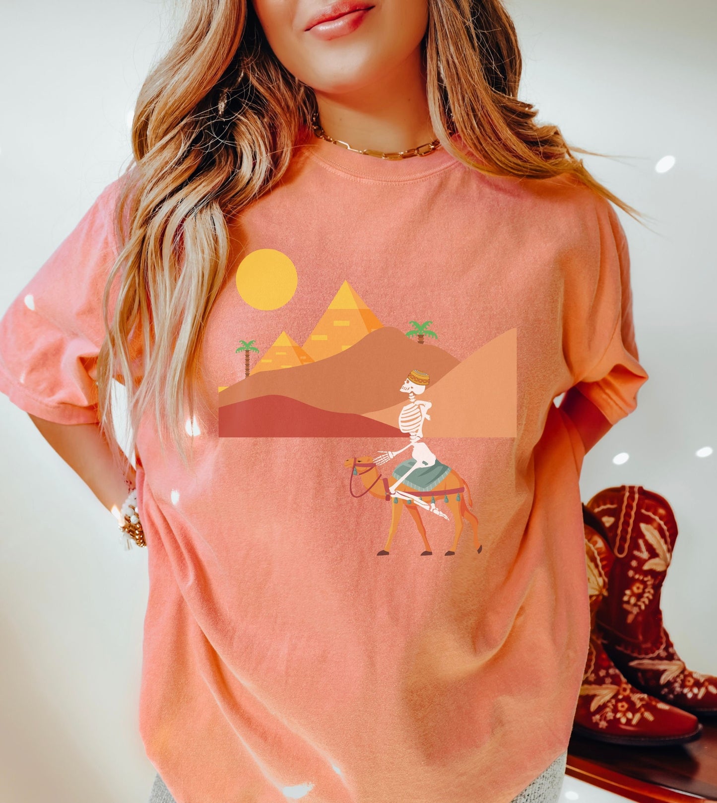 A woman wearing a cute, vintage terracotta colored shirt with a skeleton wearing an Egyptian hat riding a brown camel through the desert, the Egyptian pyramids in the distance behind sand dunes and palm trees with the sun high in the sky.