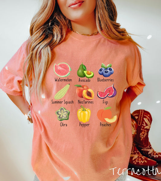 A woman wearing a vintage, terracotta colored shirt with the picture and text of a watermelon, an avocado, blueberries, a summer squash, nectarines, figs, oka, a pepper, and peaches listed in three rows of three.