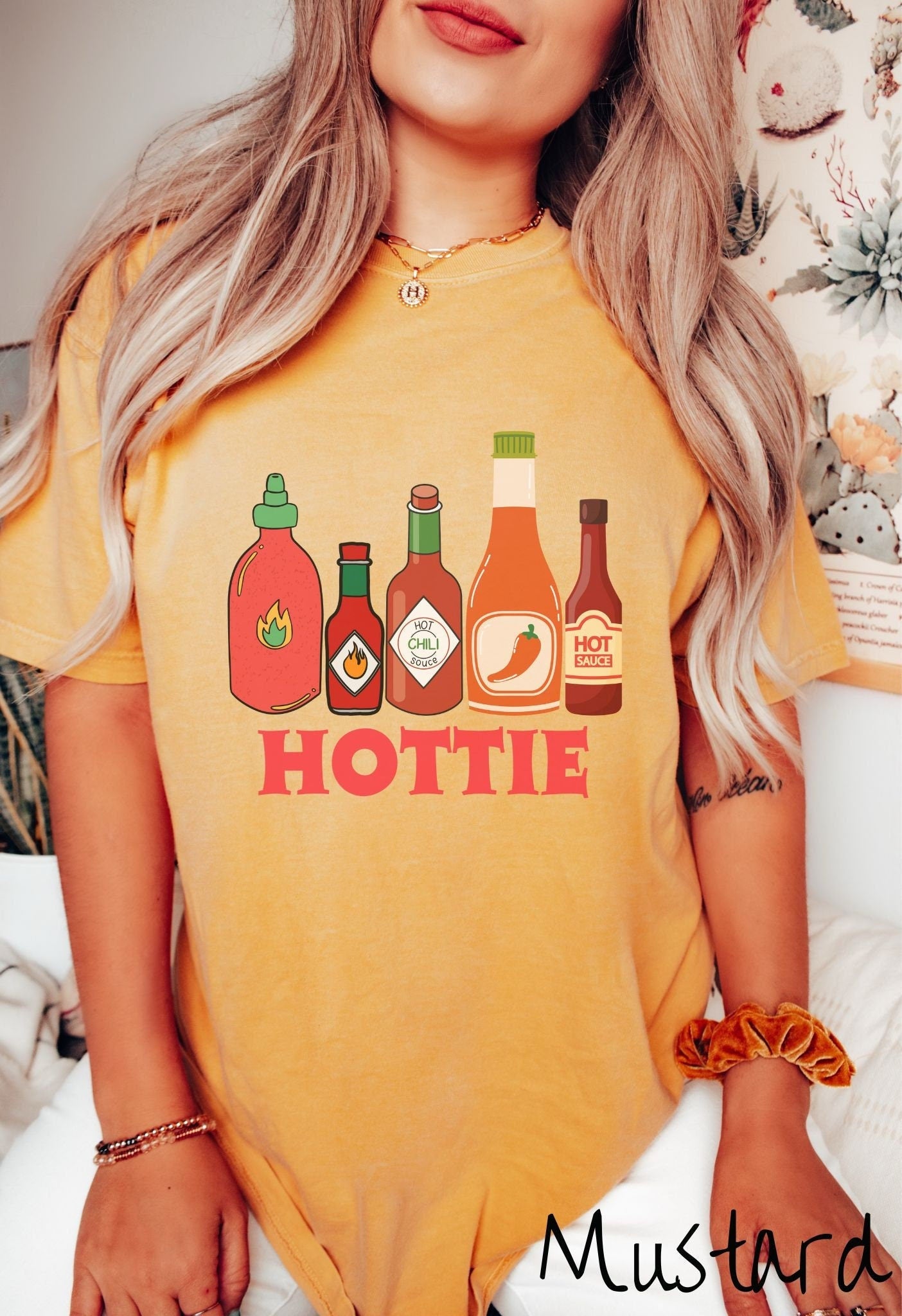 A woman wearing a vintage, mustard colored shirt with red, orange, and dark red bottles of spicy and hot sauces with fire and hot pepper pictures on the labels and the text Hottie in thick red font underneath the bottles and centered on the shirt.