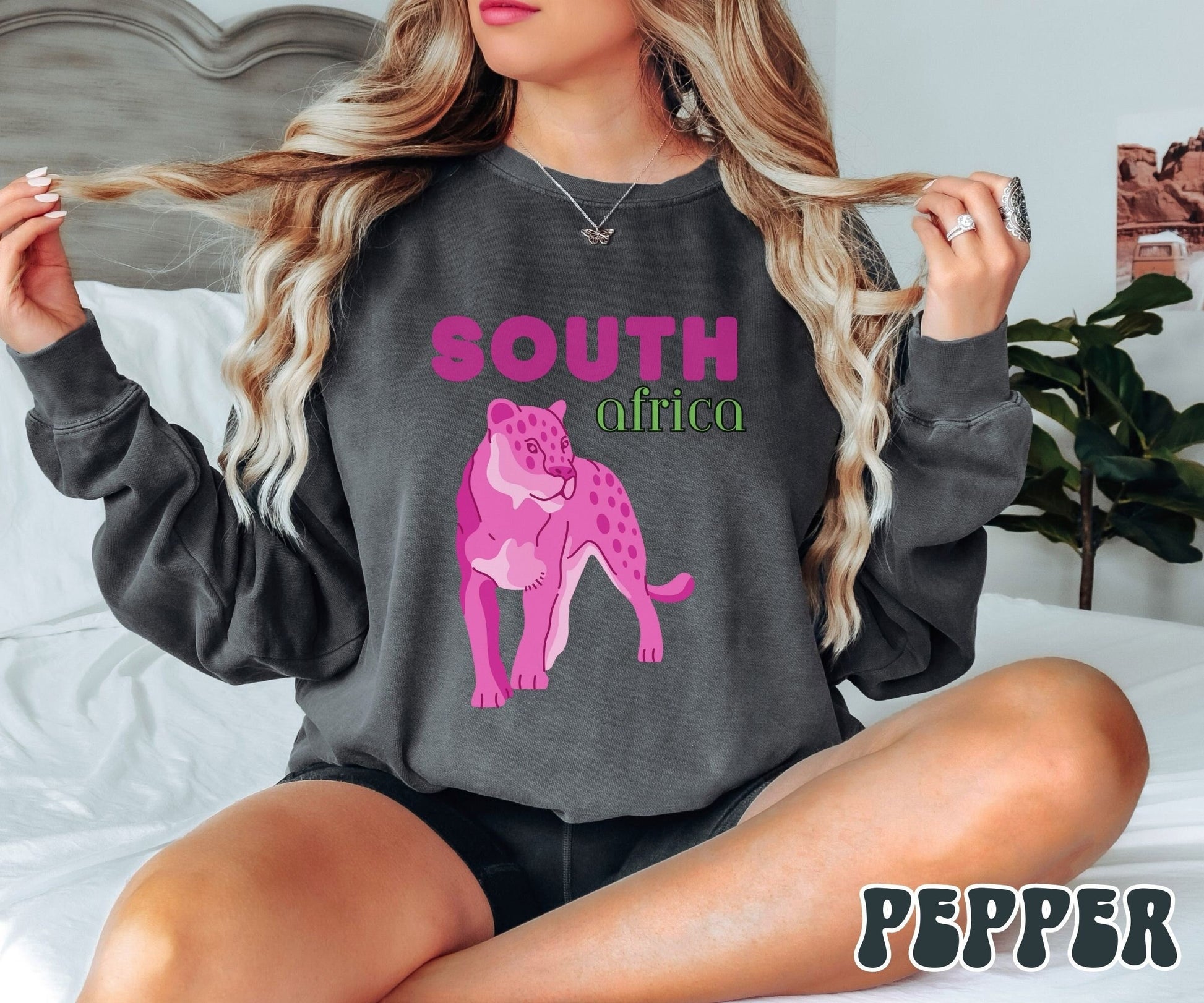 A woman wearing a vintage, pepper colored sweatshirt with the text South Africa in pink and green font, respectively, and a pink tiger underneath on the prowl.
