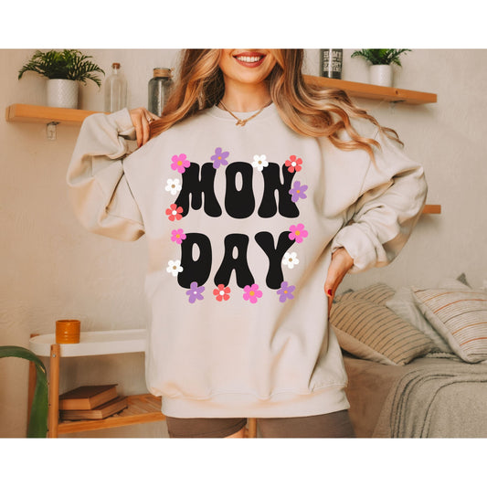 A woman wearing a vintage, sand colored sweatshirt with the text centered on the front MON then DAY below it, with pink, purple, white, and orange flowers scattered amongst the letters.