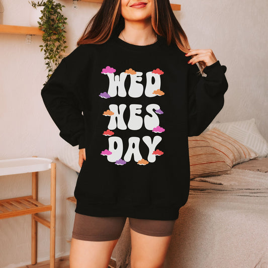 A woman wearing a vintage, black colored sweatshirt with the text centered on the front WED then below it NES then DAY below that, with pink, red, purple, and orange clouds scattered amongst the letters.