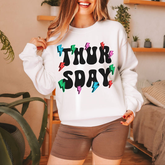 A woman wearing a vintage, white colored sweatshirt with the text centered on the front THURS then below it DAY, with pink, red, blue, and green lightning bolts scattered amongst the letters.