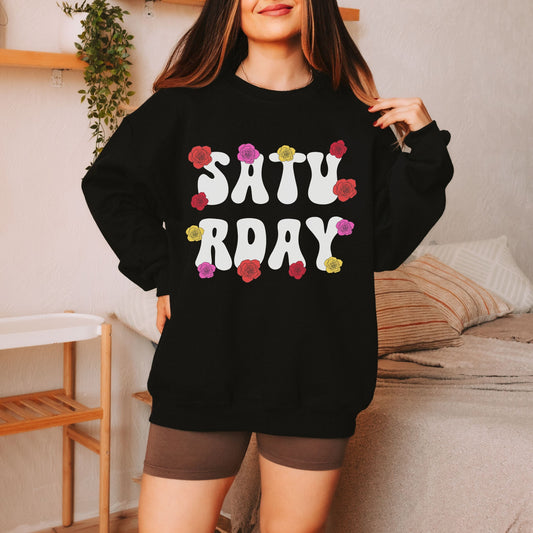 A woman wearing a vintage, black colored sweatshirt with the text centered on the front SATU then below it RDAY, with pink, red, and yellow roses scattered amongst the letters.