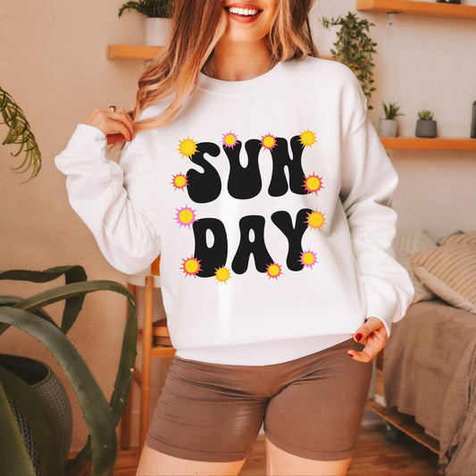 A woman wearing a vintage, white colored sweatshirt with the text centered on the front SUN then below it DAY, with yellow, orange, and pink suns scattered amongst the letters.