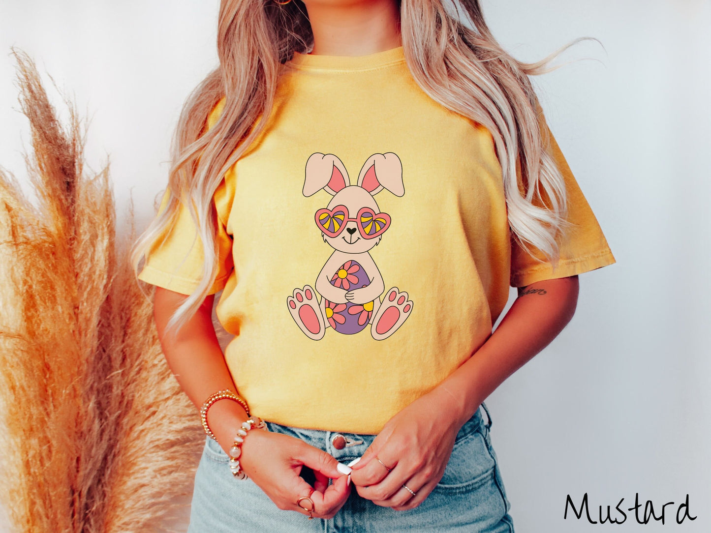 A woman wearing a cute, vintage mustard colored shirt with a smiling ivory and pink colored rabbit holding a large purple Easter egg with pink flowers. The rabbit is wearing heart-shaped pink glasses and has a heart-shaped nose.