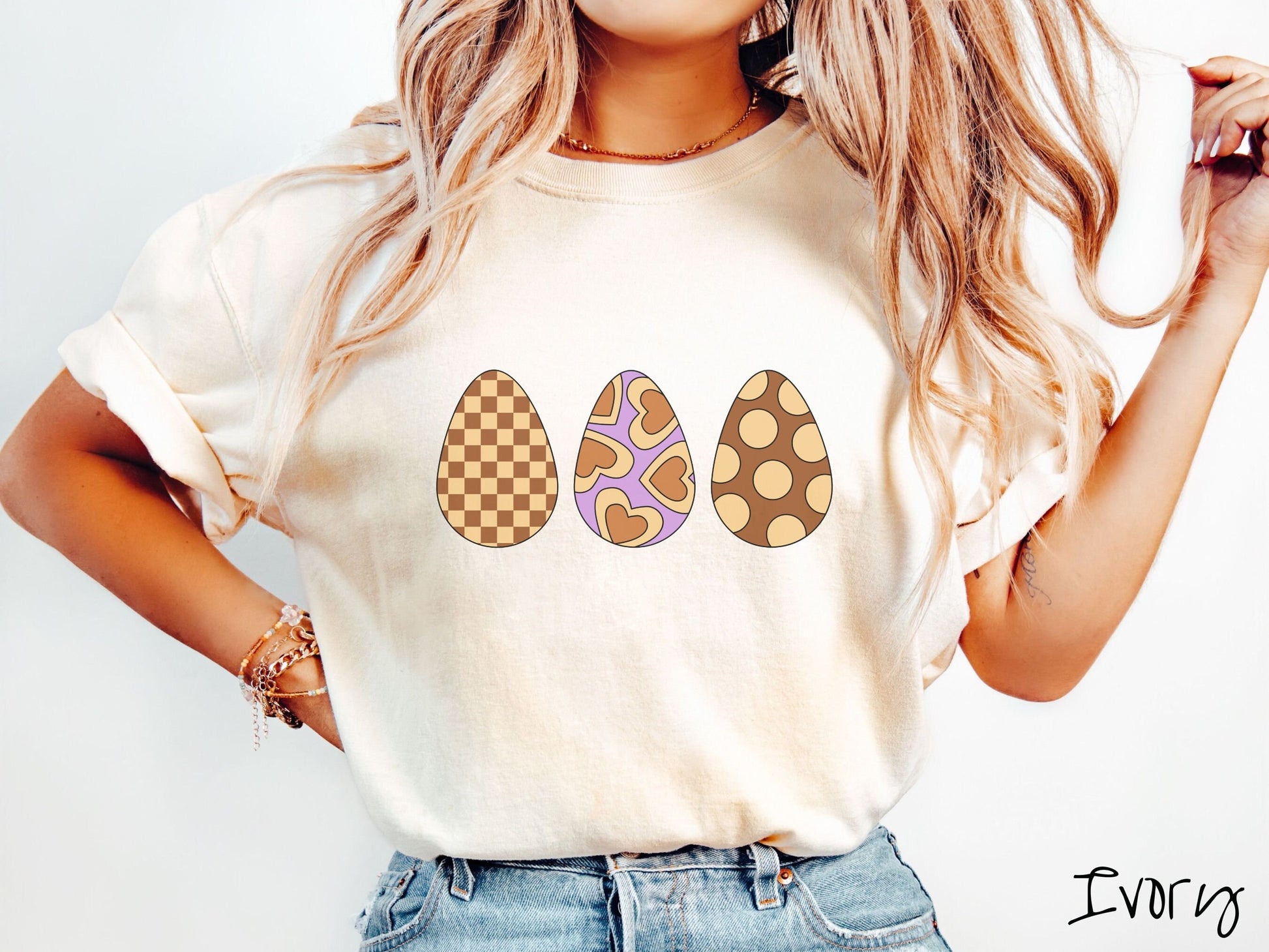 A woman wearing a cute, vintage ivory colored shirt with three Easter eggs. The left egg has a light yellow and brown checkerboard pattern, the middle is purple with light yellow brown hearts, and the right is brown with light yellow circles.