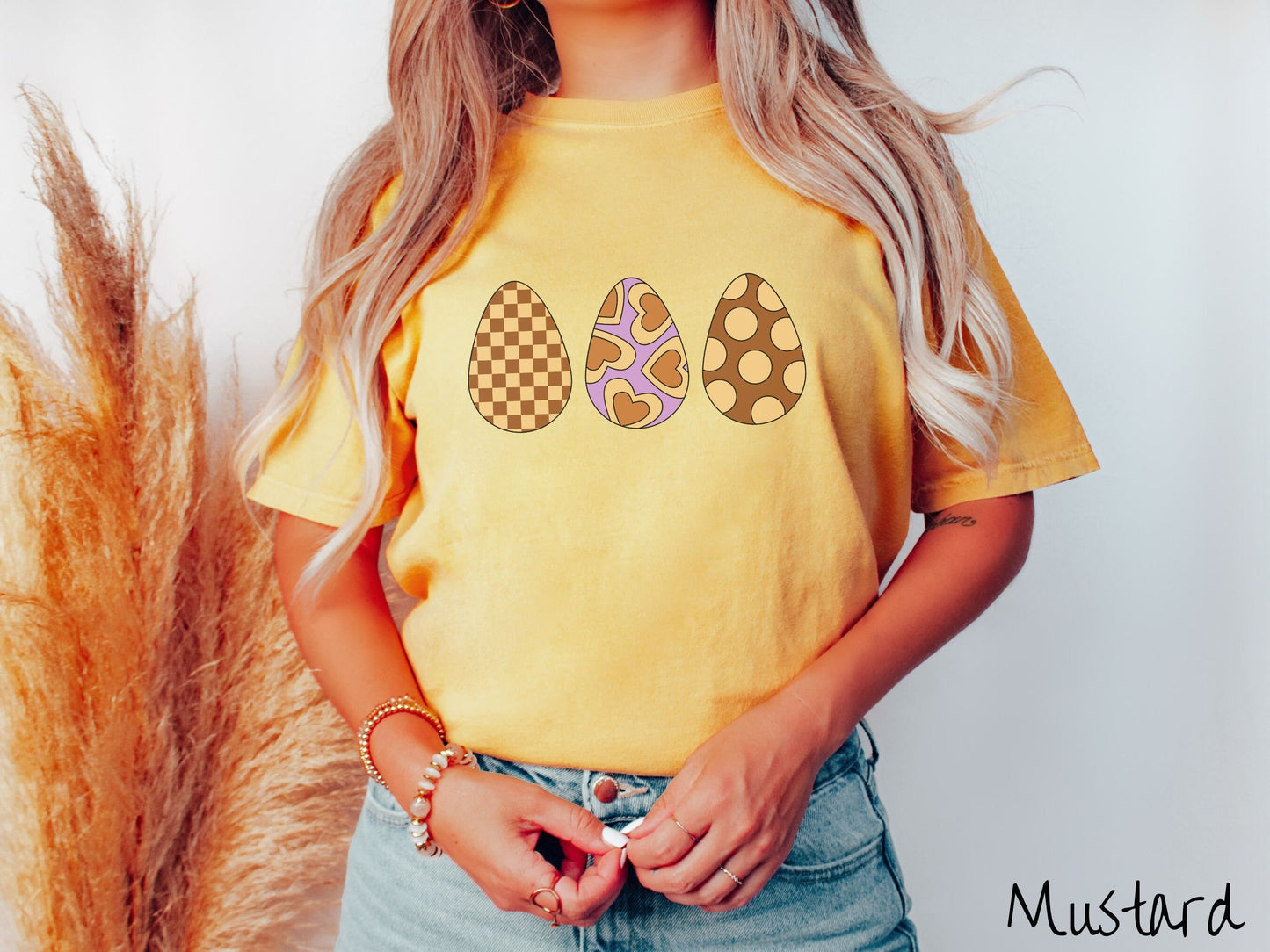 A woman wearing a cute, vintage mustard colored shirt with three Easter eggs. The left egg has a light yellow and brown checkerboard pattern, the middle is purple with light yellow brown hearts, and the right is brown with light yellow circles.