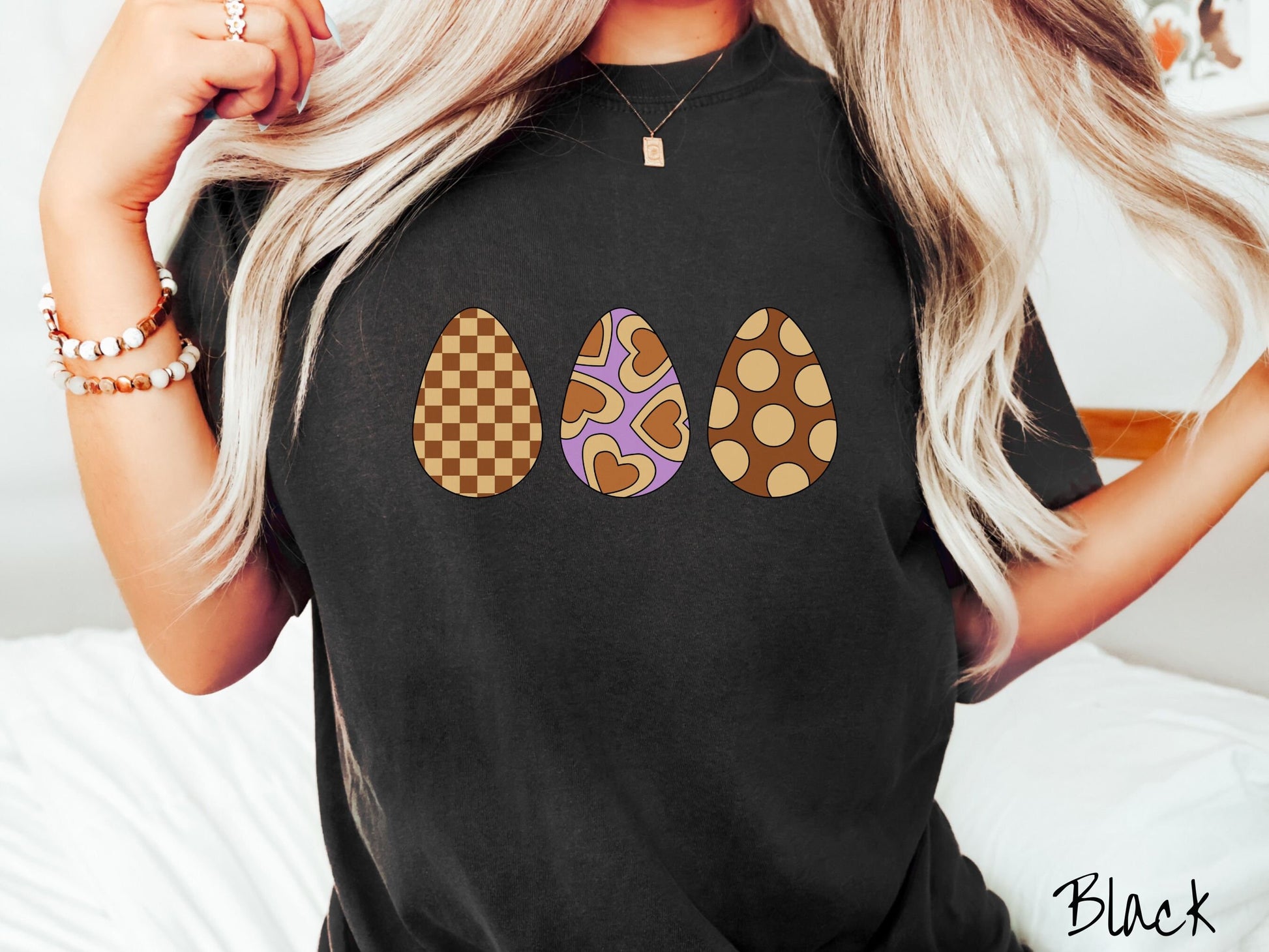A woman wearing a cute, vintage black colored shirt with three Easter eggs. The left egg has a light yellow and brown checkerboard pattern, the middle is purple with light yellow brown hearts, and the right is brown with light yellow circles.