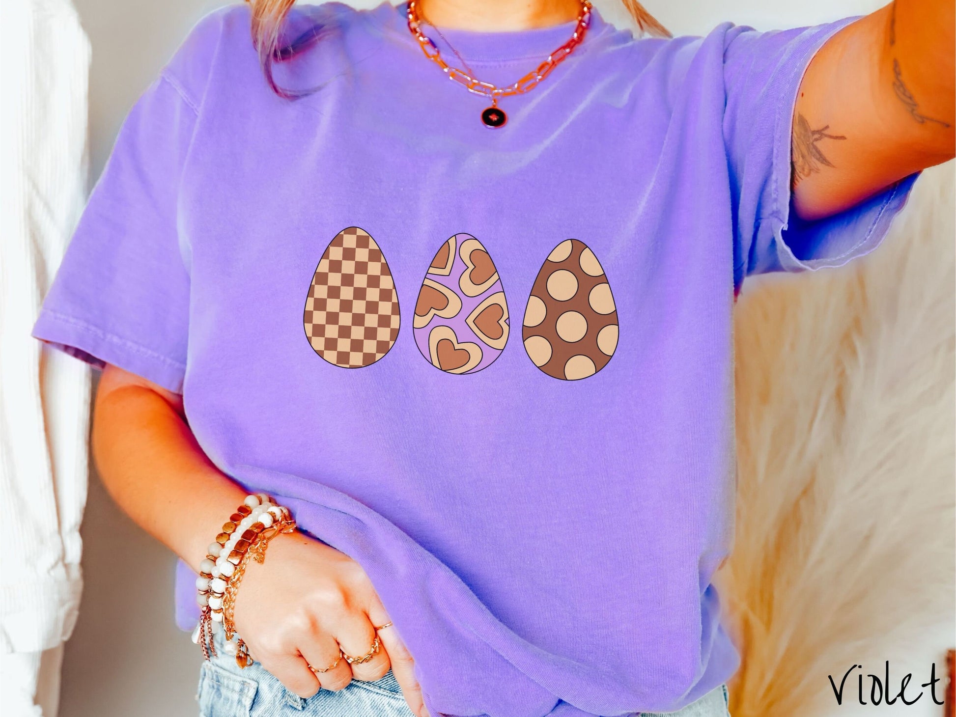 A woman wearing a cute, vintage violet colored shirt with three Easter eggs. The left egg has a light yellow and brown checkerboard pattern, the middle is purple with light yellow brown hearts, and the right is brown with light yellow circles.