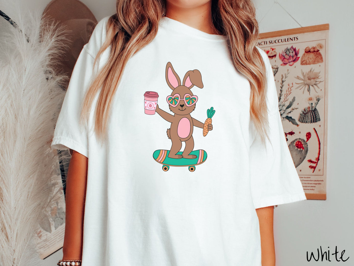 A woman wearing a cute, vintage white colored shirt with a brown bunny rabbit with heart-shaped glasses, a carrot in one hand, and a pink to-go coffee in the other, riding a green, pink, and orange skateboard.