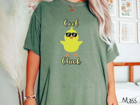 A woman wearing a cute, vintage moss colored shirt with the yellow text Cool Chick and below is a yellow baby chick with black sunglasses with its wings spread. It has an orange mouth and feet and yellow feathers sticking out the top of its head.