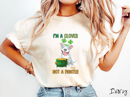 A woman wearing a vintage, ivory colored shirt with text Im a clover not a fighter in green and yellow font. In between the text is a smiling pitbull puppy wearing a hat with  two green leaf clovers, its paw resting on top of a green pot of gold.