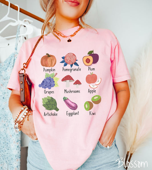 A woman wearing a vintage, blossom colored shirt with the picture and text of a pumpkin, a pomegranate, a plum, grapes, mushrooms, an apple, an artichoke, an eggplant, and a kiwi listed in three rows of three.