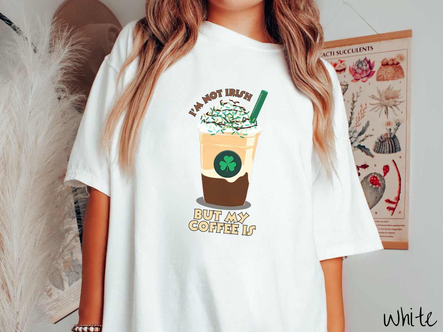 A woman wearing a vintage, white colored shirt with the text Im not Irish but my coffee is in brown and yellow text, between the text is a green and yellow coffee cup with a green clover and  whipped cream and chocolate drizzle on top.