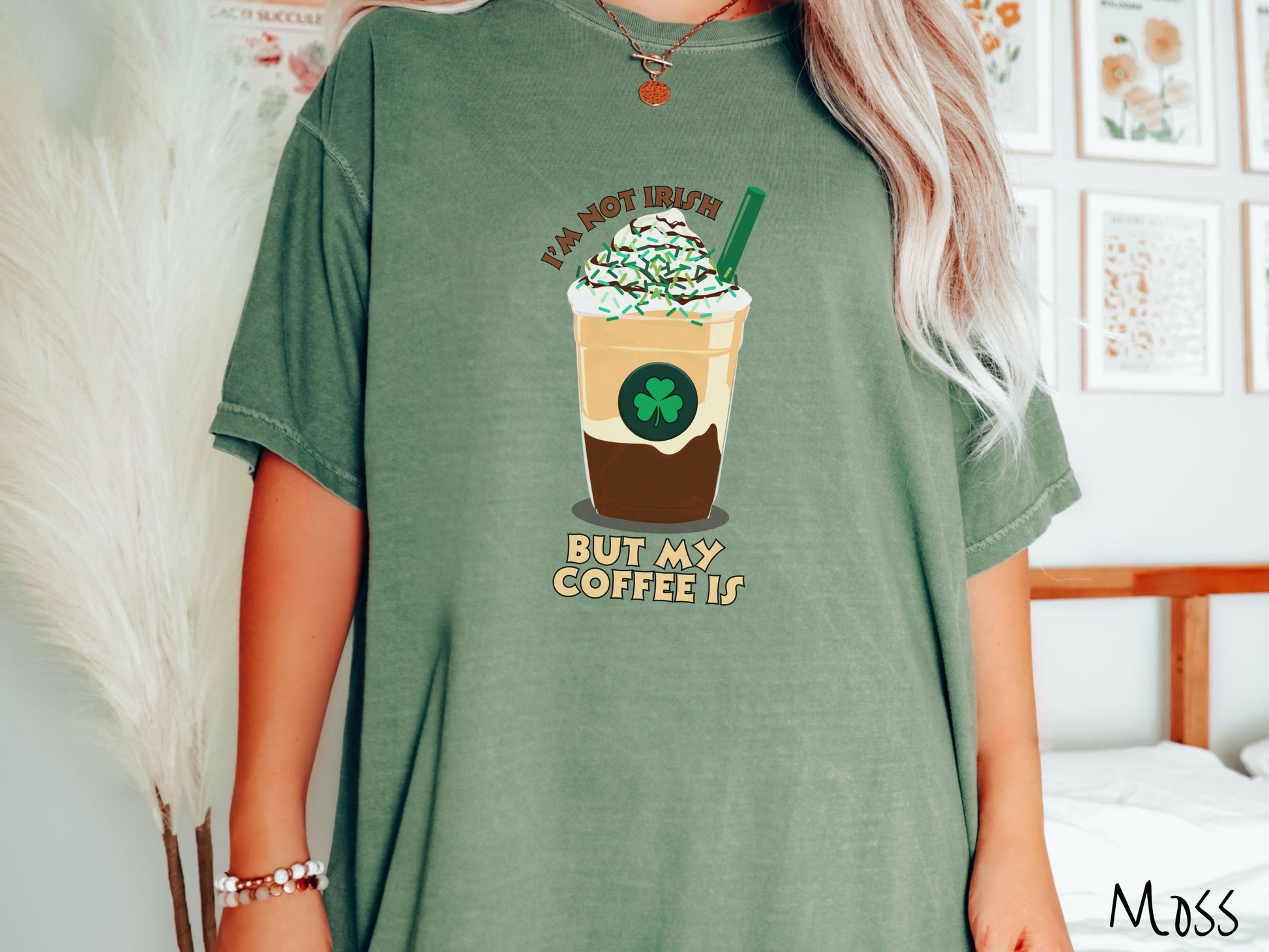 A woman wearing a vintage, moss colored shirt with the text Im not Irish but my coffee is in brown and yellow text, between the text is a green and yellow coffee cup with a green clover and  whipped cream and chocolate drizzle on top.