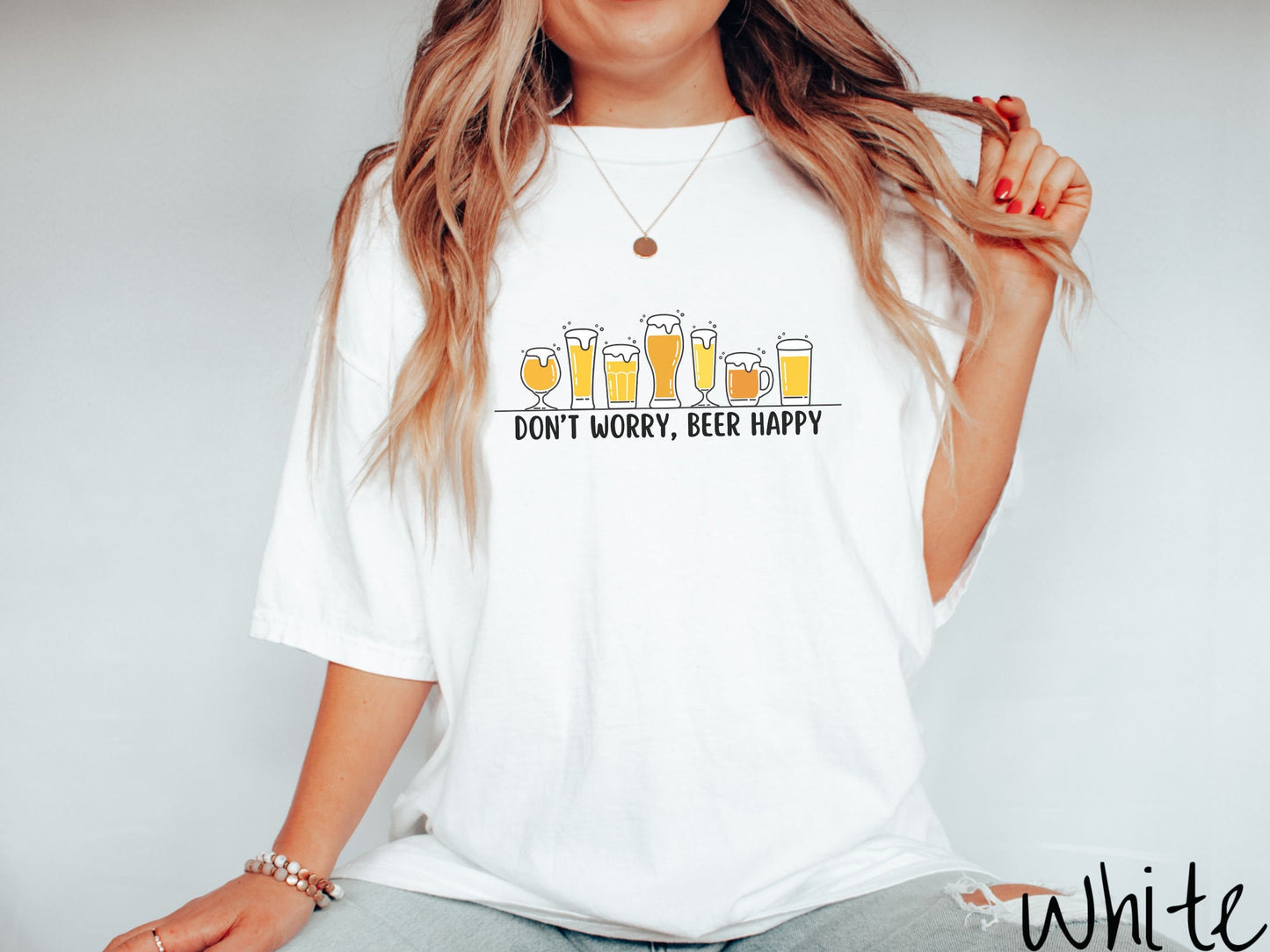 A woman wearing a vintage, white colored shirt with the text Dont Worry, Beer happy, and above this are varying sizes and shapes of beer glasses with yellow and orange ber all of them have foam coming out the tops.