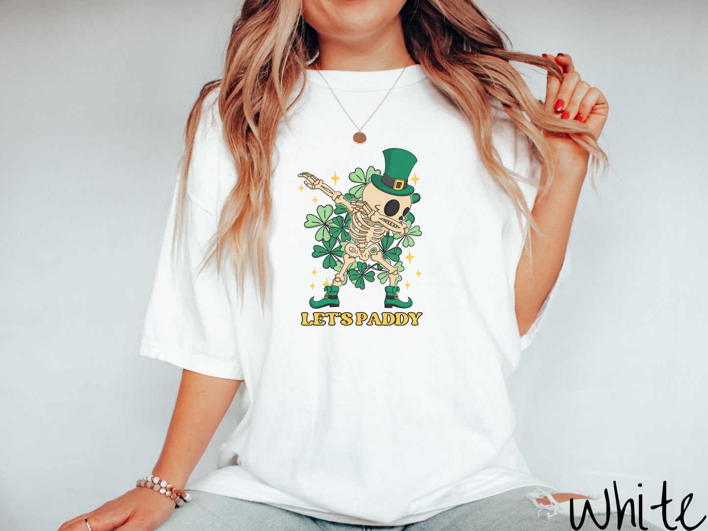 A woman wearing a vintage, white colored shirt with the text Lets Paddy and below that is a leprechaun wearing a green top hat and green elf shoes doing the dab dance and there are green clovers and golden stars in the background.
