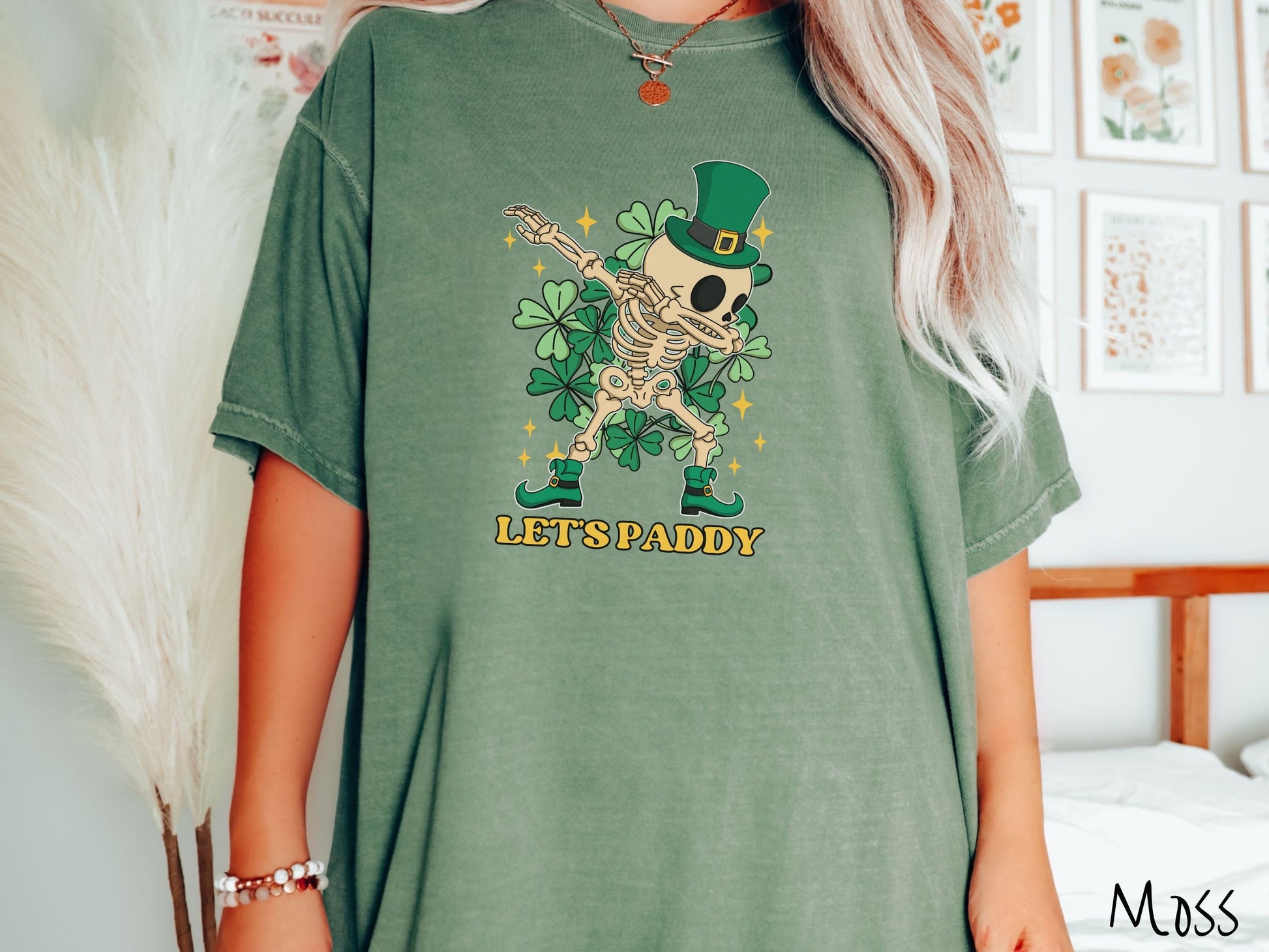 A woman wearing a vintage, moss colored shirt with the text Lets Paddy and below that is a leprechaun wearing a green top hat and green elf shoes doing the dab dance and there are green clovers and golden stars in the background.