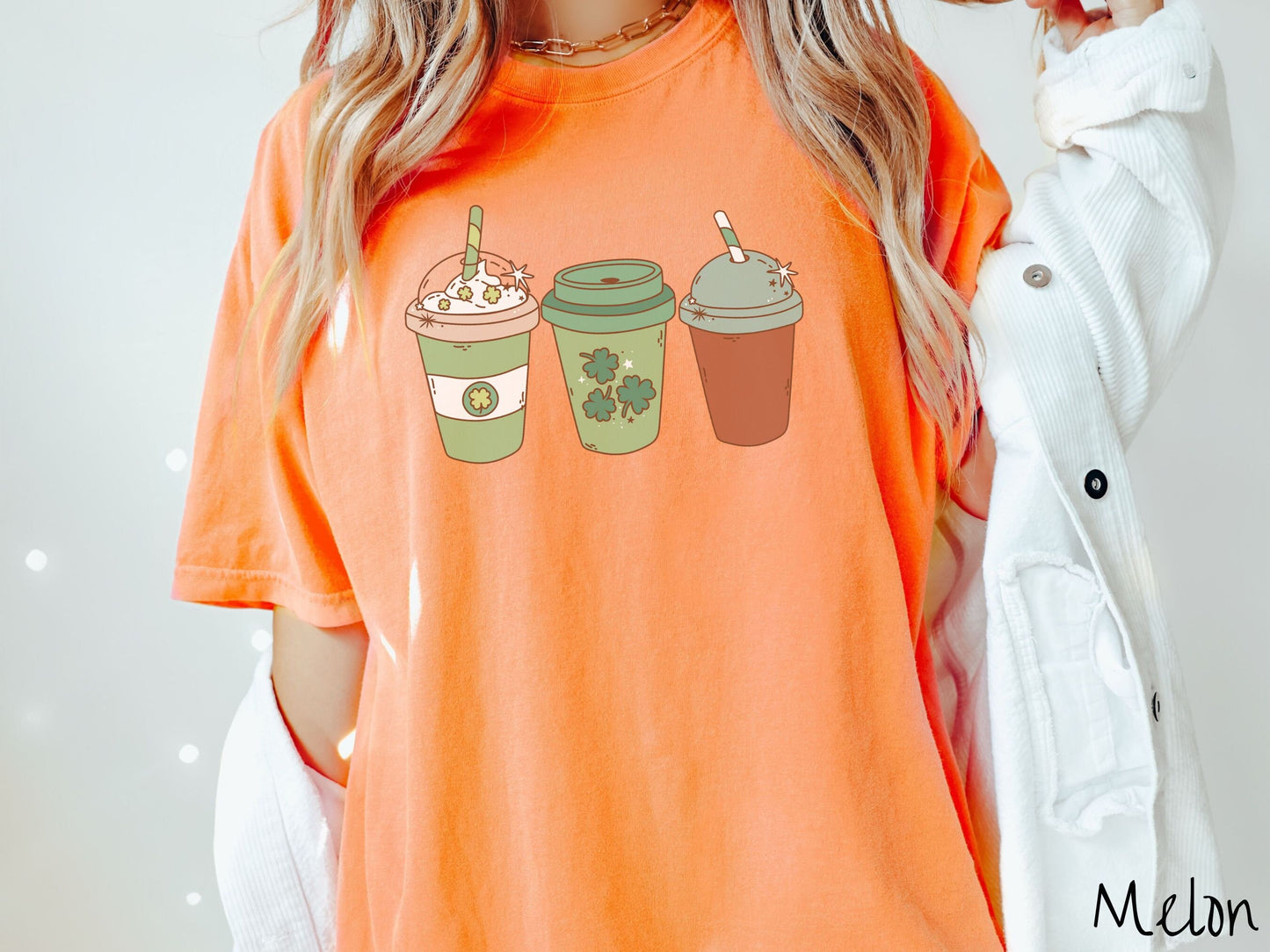 A woman wearing a vintage, melon colored shirt with three coffee cups, one is white and green with a clear lid showing whipped cream topping with green clover sprinkles, another green cup with green clovers, and the third is brown with a green lid.