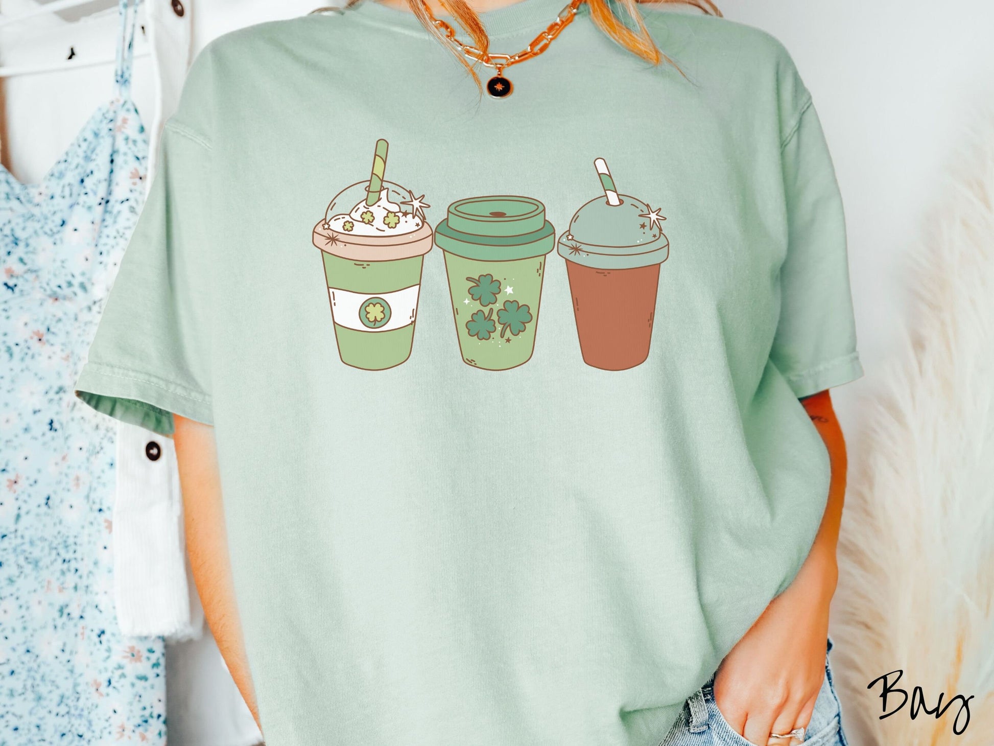 A woman wearing a vintage, bay colored shirt with three coffee cups, one is white and green with a clear lid showing whipped cream topping with green clover sprinkles, another green cup with green clovers, and the third is brown with a green lid.