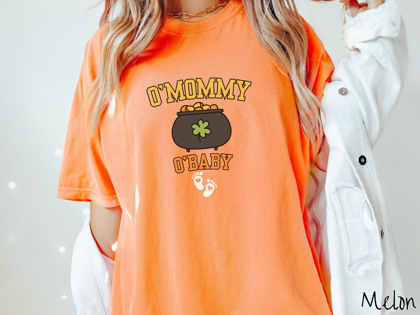 A woman wearing a vintage, melon colored shirt with text O Mommy O Baby in yellow and orange font, a black pot of gold with a green clover on it and yellow and orange gold coins inside. Below all this are two white baby footprints with pink hearts.