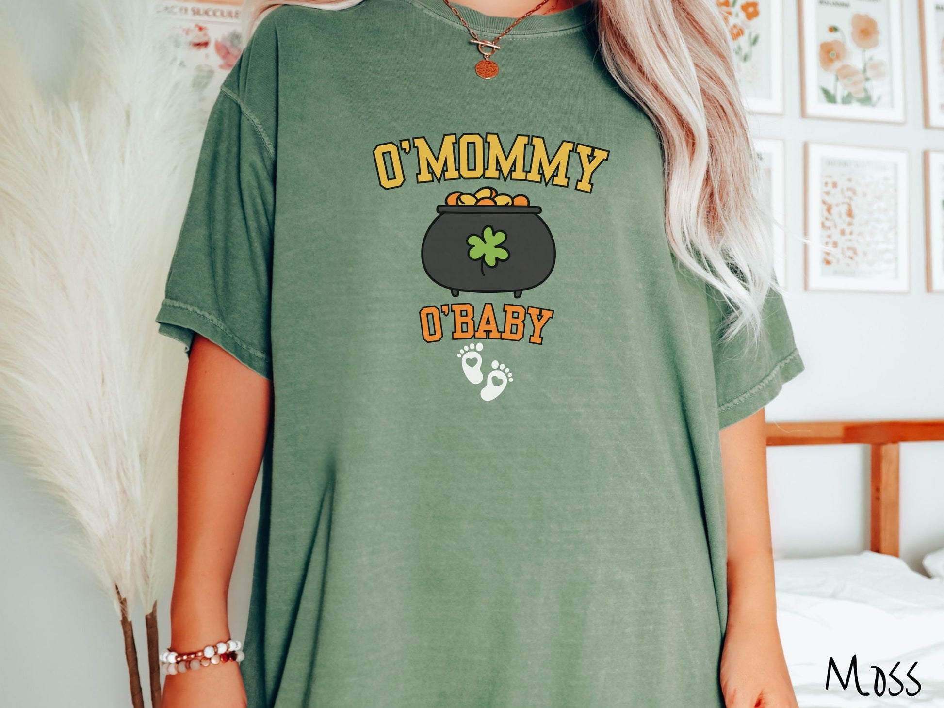 A woman wearing a vintage, moss colored shirt with text O Mommy O Baby in yellow and orange font, a black pot of gold with a green clover on it and yellow and orange gold coins inside. Below all this are two white baby footprints with pink hearts.