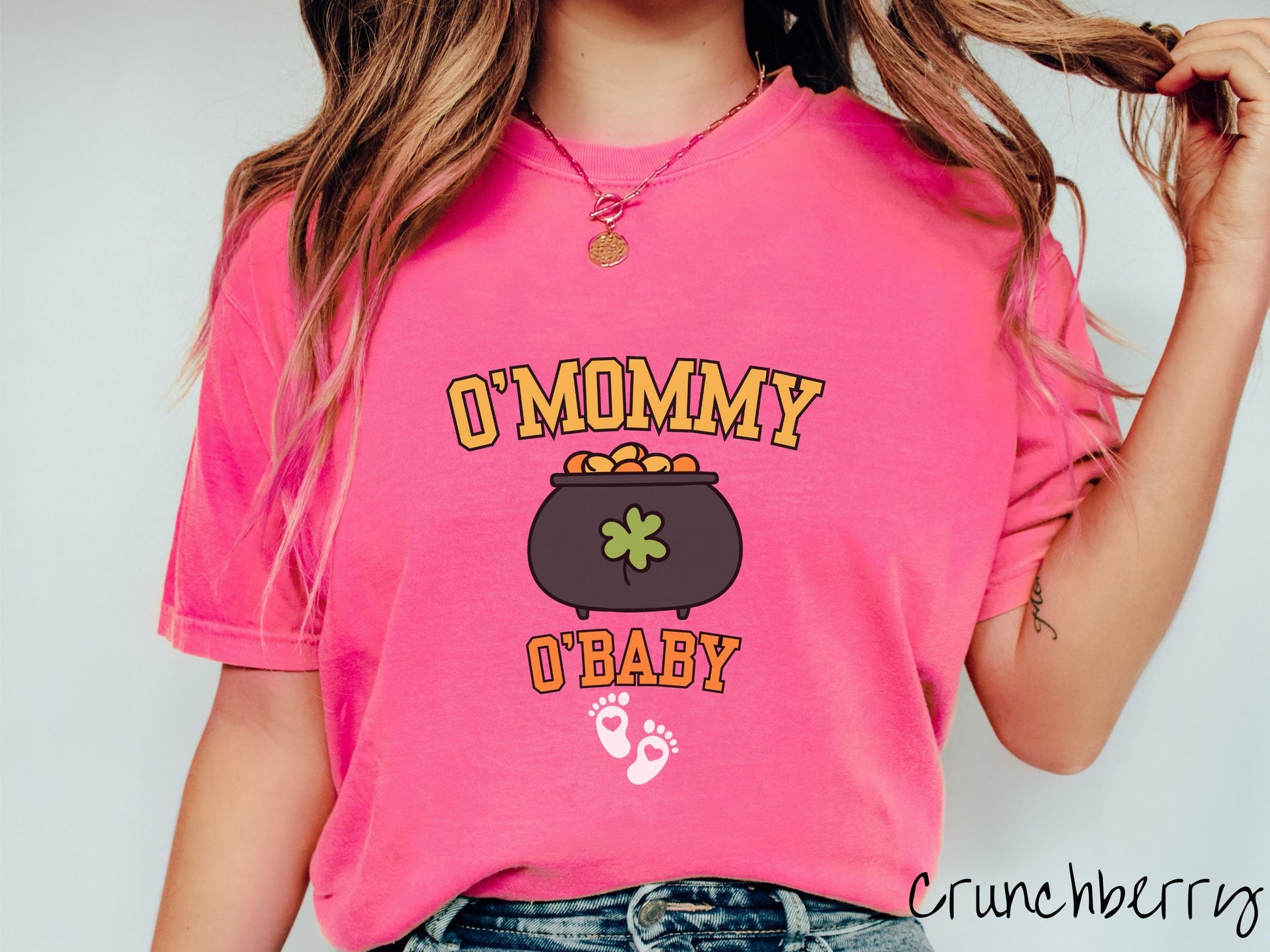 A woman wearing a vintage, crunchberry colored shirt with text O Mommy O Baby in yellow and orange font, a pot of gold with a green clover on it and yellow and orange gold coins inside. Below all this are two white baby footprints with pink hearts.