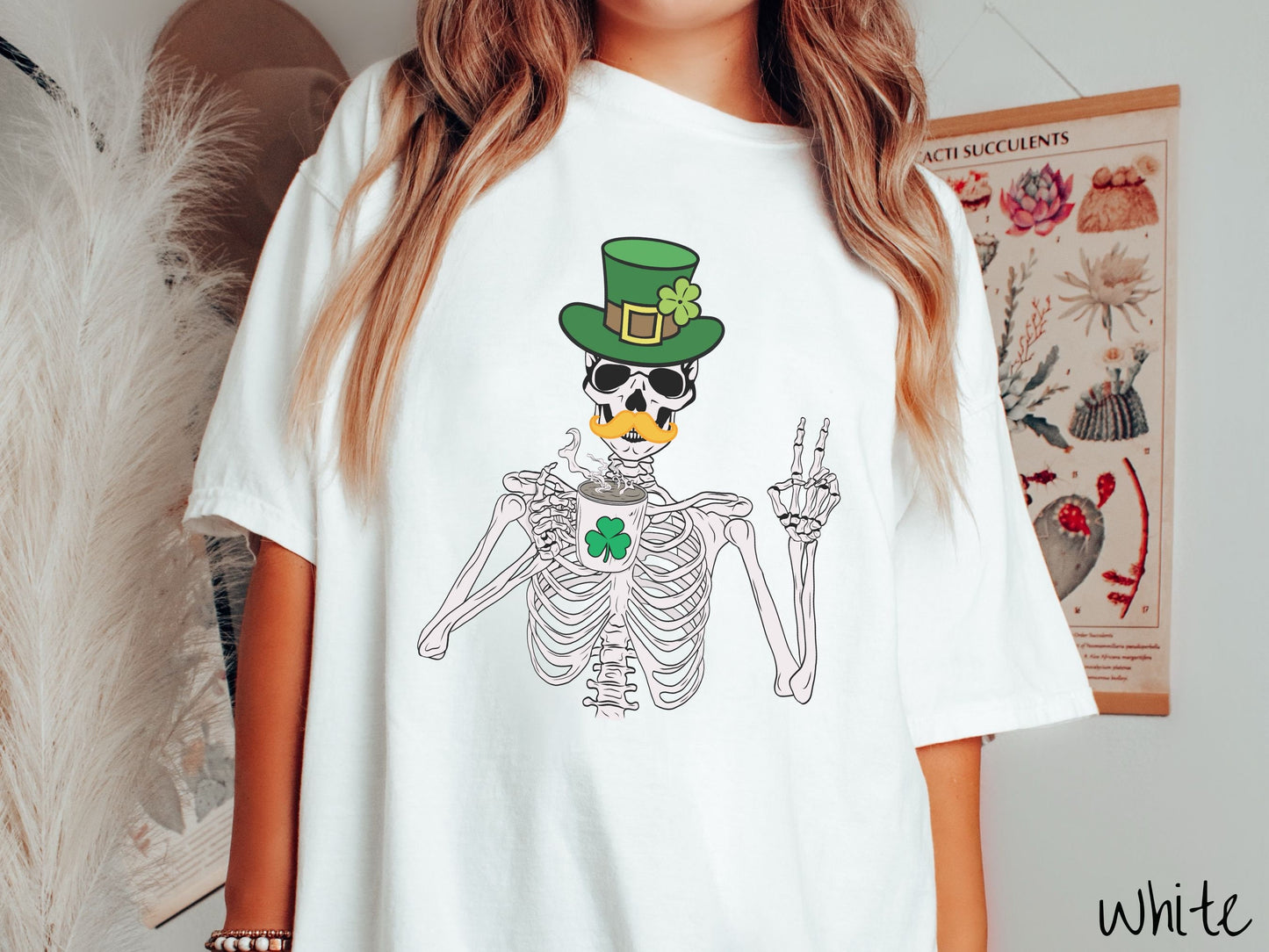 A woman wearing a vintage, white colored shirt with a skeleton wearing a green top hat with a green clover and an orange mustache showing the peace sign with its left hand and drinking out of a steaming hot coffee cup with a three leaf clover on it.