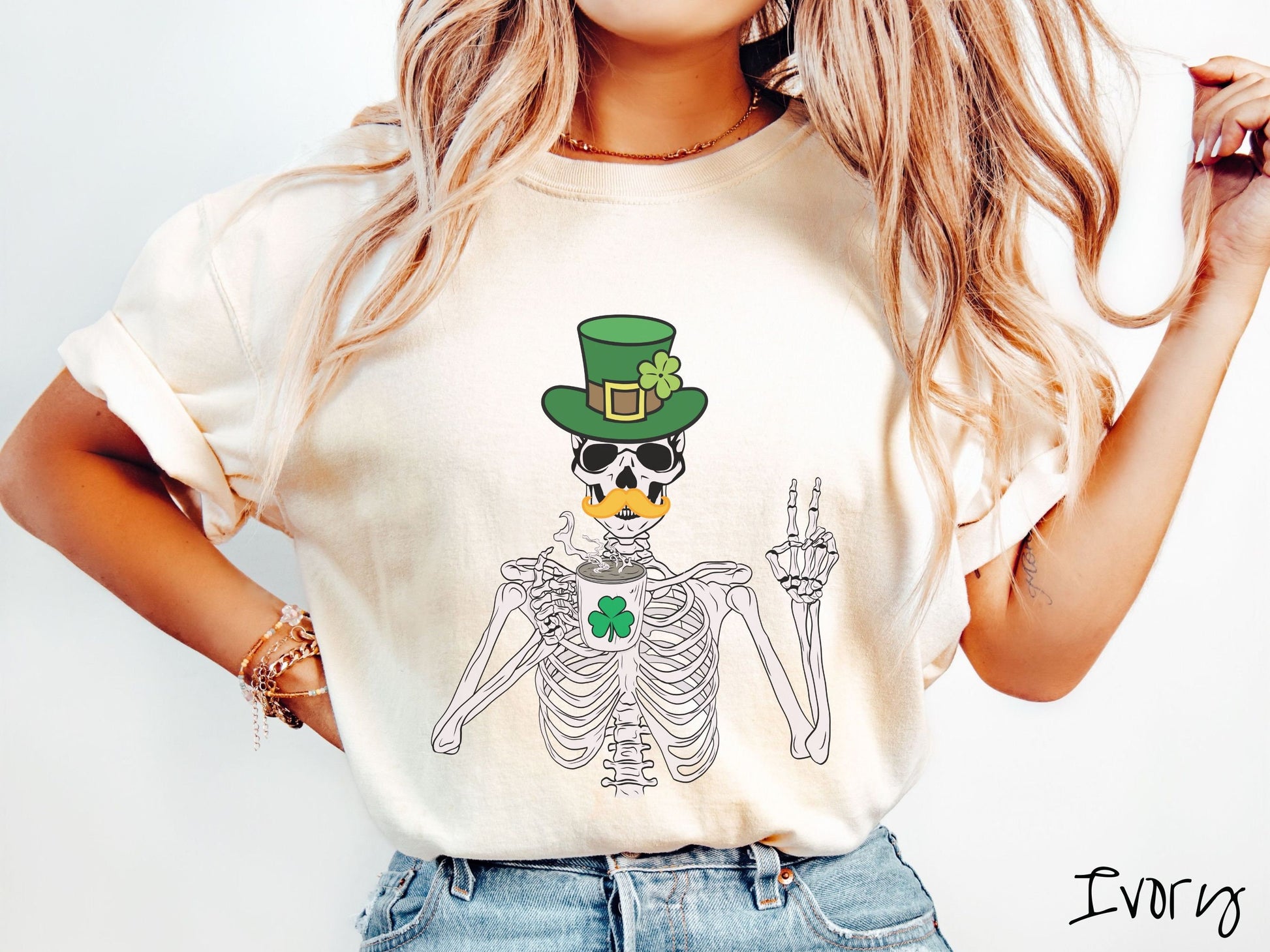 A woman wearing a vintage, ivory colored shirt with a skeleton wearing a green top hat with a green clover and an orange mustache showing the peace sign with its left hand and drinking out of a steaming hot coffee cup with a three leaf clover on it.