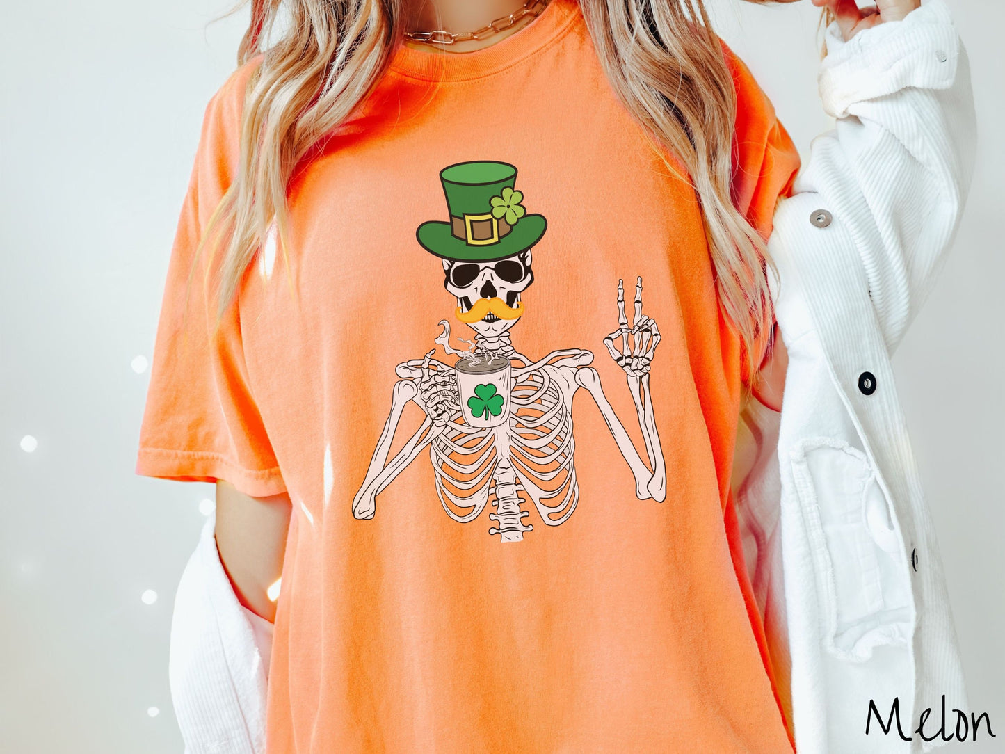 A woman wearing a vintage, melon colored shirt with a skeleton wearing a green top hat with a green clover and an orange mustache showing the peace sign with its left hand and drinking out of a steaming hot coffee cup with a three leaf clover on it.