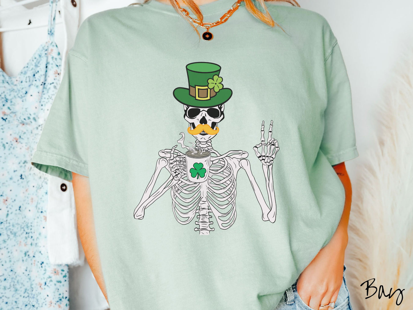 A woman wearing a vintage, bay colored shirt with a skeleton wearing a green top hat with a green clover and an orange mustache showing the peace sign with its left hand and drinking out of a steaming hot coffee cup with a three leaf clover on it.